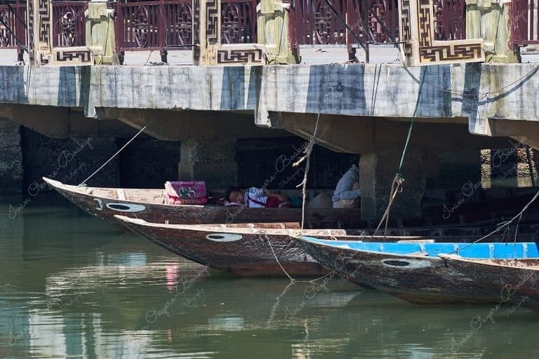 Young girl in school clothes having an afternoon nap in her mother’s boat in the shade underneath the Bridge of Lights (Cau An Hoi) in Hoi An, Vietnam