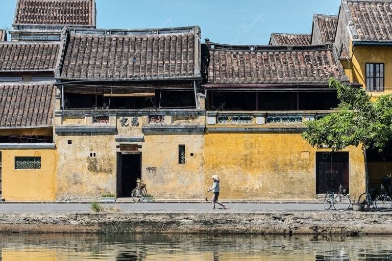 View of a Vietnamese woman wearing a traditional non la hat walking in front of a classic yellow pastel coloured house with weathered walls at Hoi Ann Old Town riverfront, Vietnam