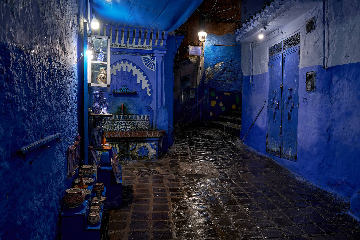 Illuminated souvenirs shop and mosaic walled drinking water fountain in an empty street at night in blue walled old town medina, Chefchaouen, Morocco