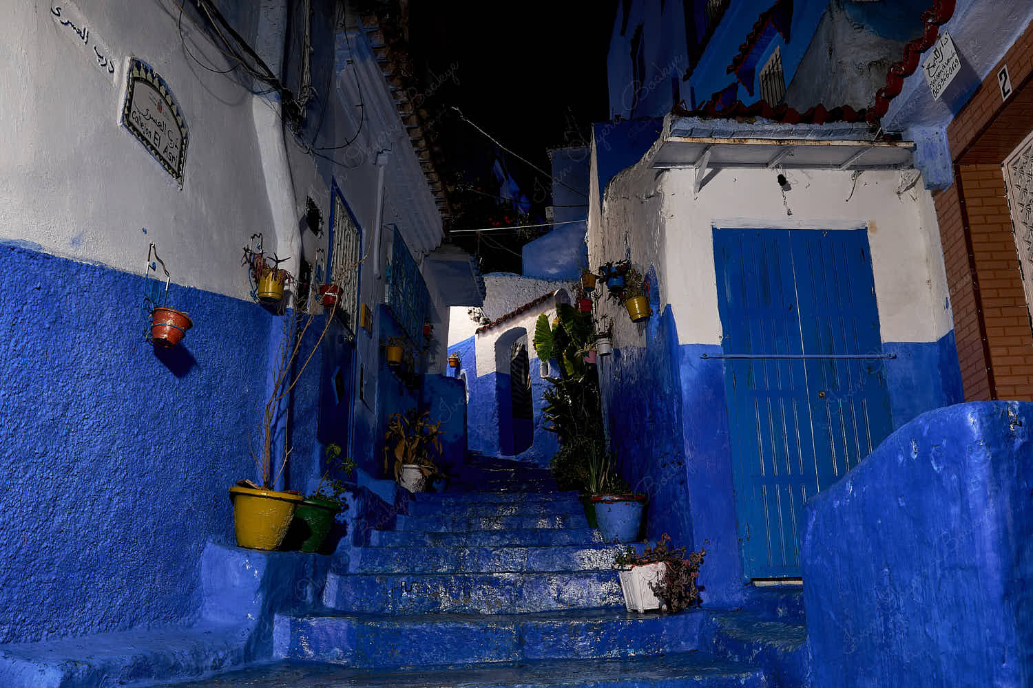 Illuminated blue steps, colorful pots and blue walls in an empty Callejon el Asri side street at night in old town medina, Chefchaouen, Morocco