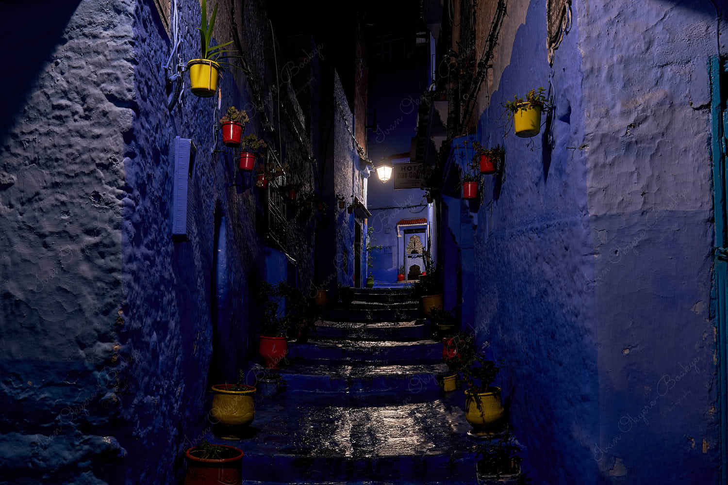 Dimly lit empty blue walled street with red and yellow flower pots and plants at night in the old town medina, Chefchaouen, Morocco
