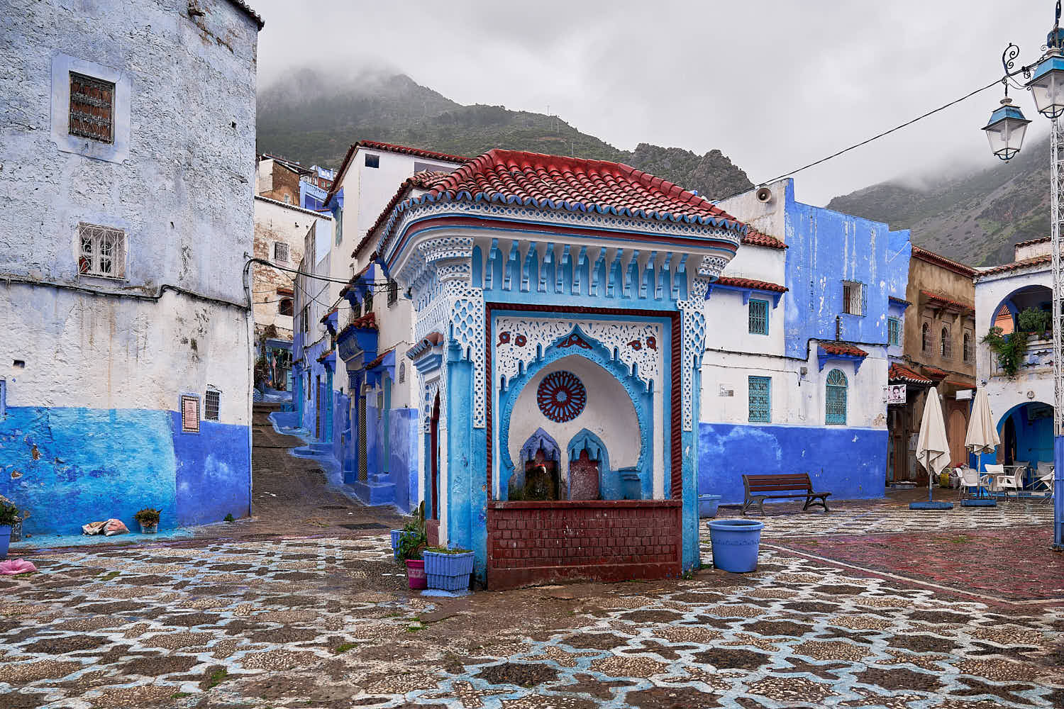 Mosaic drinking water fountain in empty Place el Haouta with clouds at the top of a mountain in the blue walled old town medina, Chefchaouen, Morocco