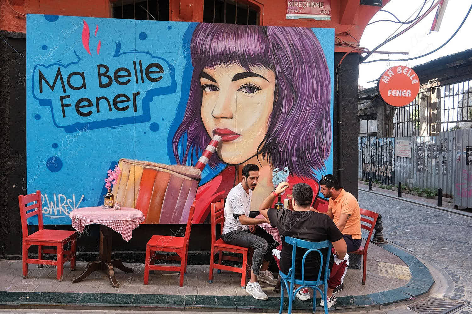 Male friends relaxing and sitting outside a colourful street mural advert on the wall of a trendy cafe in Balat, Istanbul, Turkey