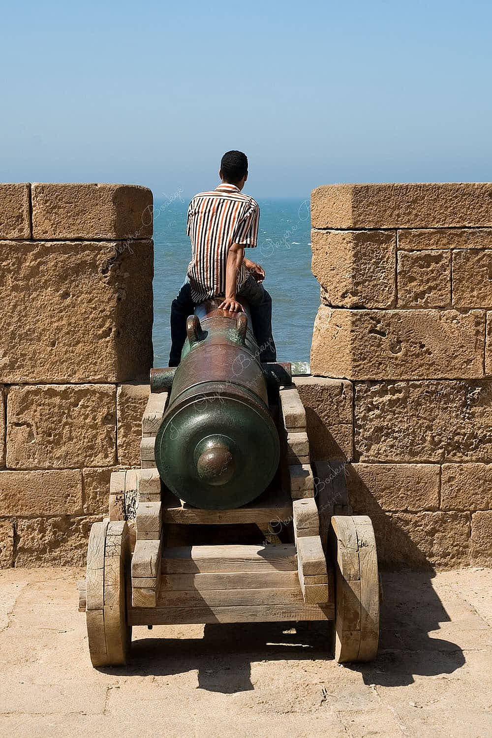 Moroccan man setting on a cannon on the ramparts of Essaouira and looking out to sea, Essaouira, Morocco