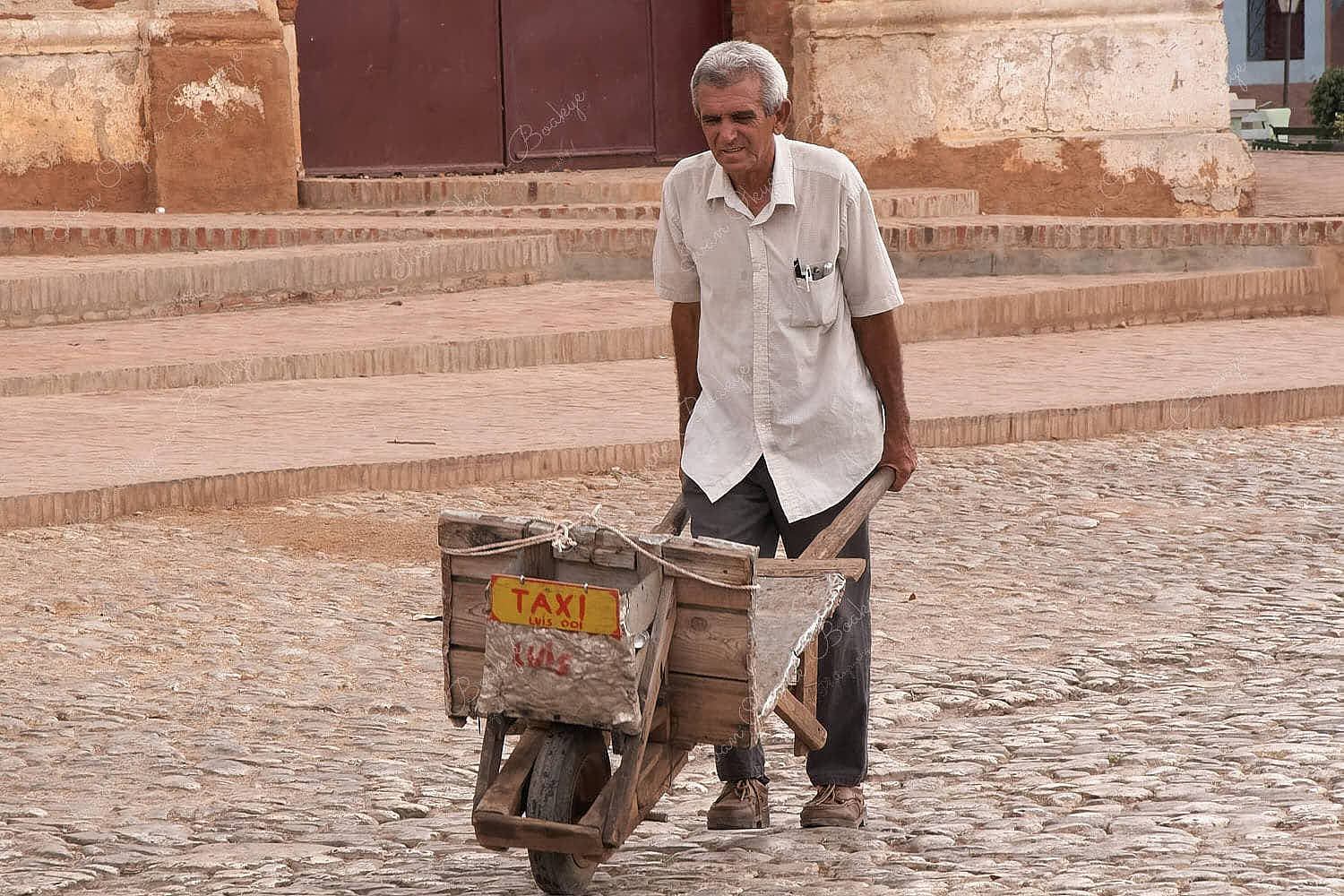 An elderly man pushing a wheelbarrow with a taxi sign attached to the front through the cobbled streets of Trinidad, Cuba