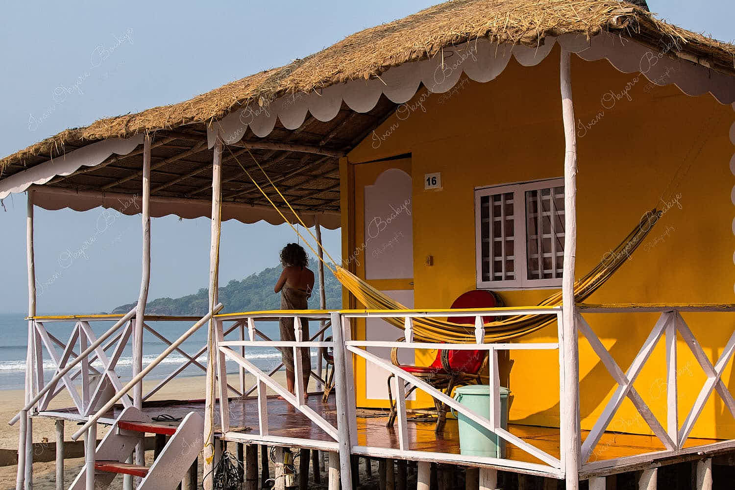 Woman standing on the porch of a bright yellow beach hut with a hammock at Palolem Beach in Goa, India