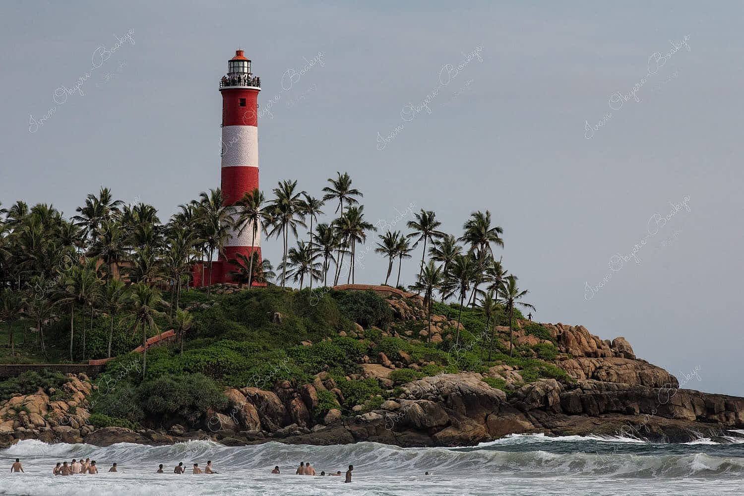 Tourists swimming at Lighthouse Beach with the Vizhinjam Lighthouse in the background in Kovalam, Kerala, India