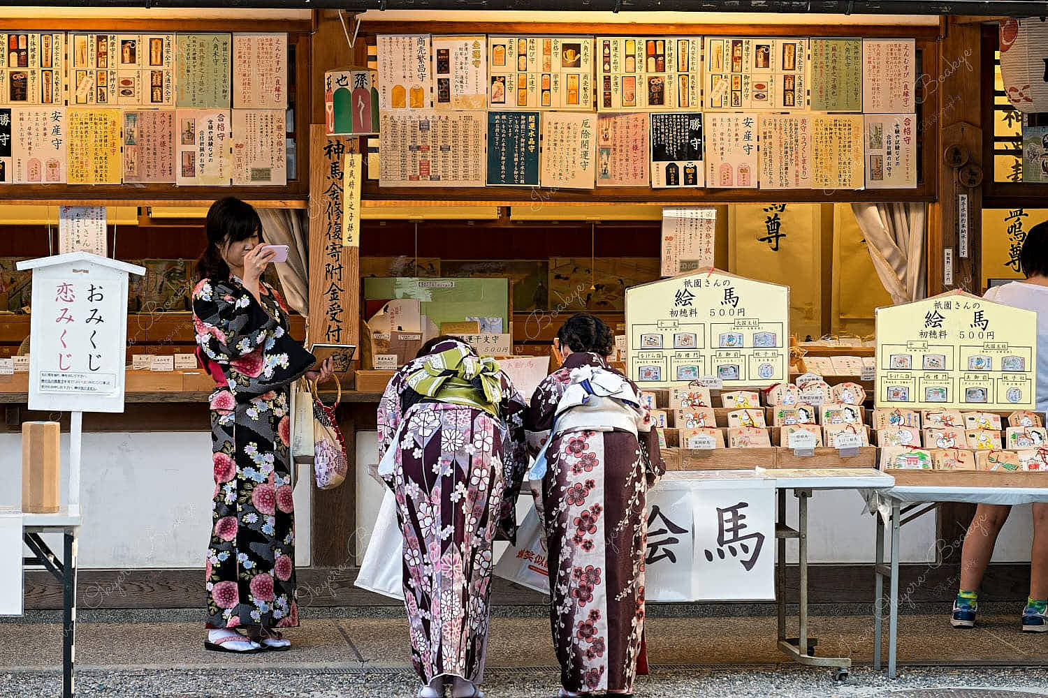 2 Japanese girlfriends wearing kimono buying ema whilst their friend takes a photo of them at Yasaka Shrine, Kyoto, Japan