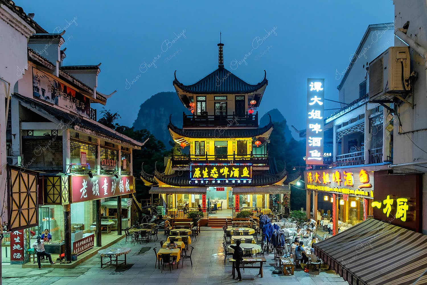 Night view of a square with table and chairs surrounded by illuminated traditional Chinese restaurants against a mountain backdrop, Yangshou, China