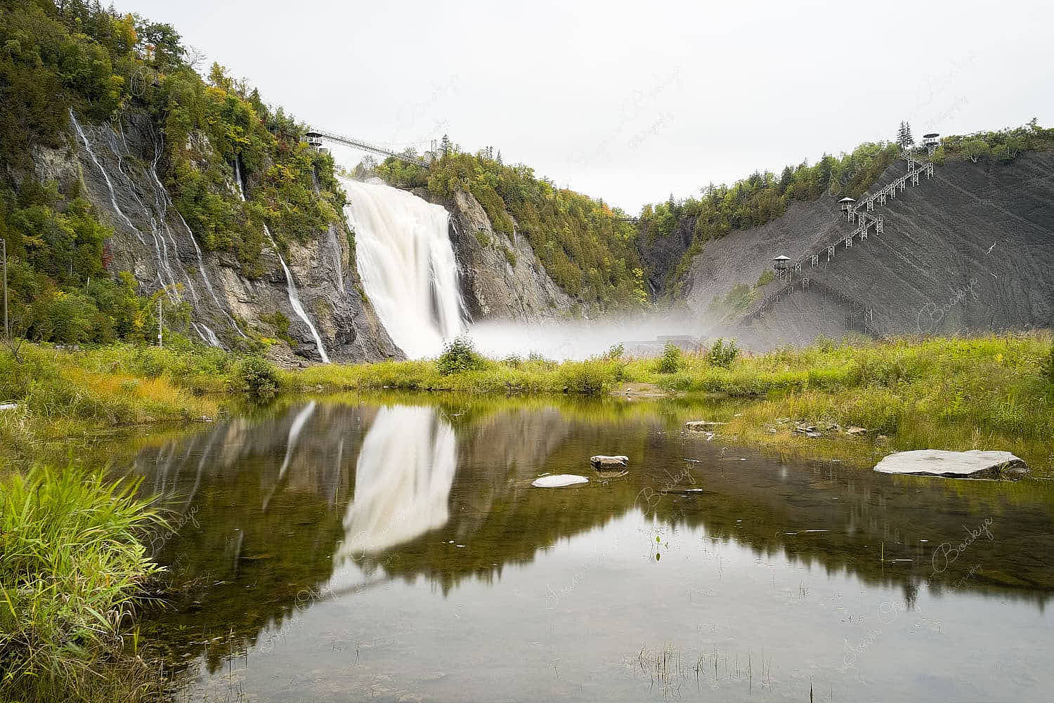 View of Montmorency Falls and Bridge and their reflected images in the surrounding water pool from a ground level, Quebec, Canada