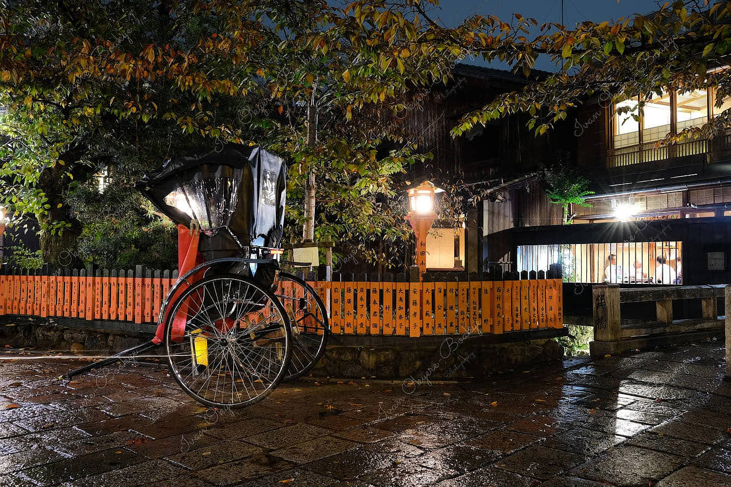 A beautiful night time view of a wet rickshaw in front of a restaurant’s vermillion orange wooden gate in a side street in Kyoto, Japan