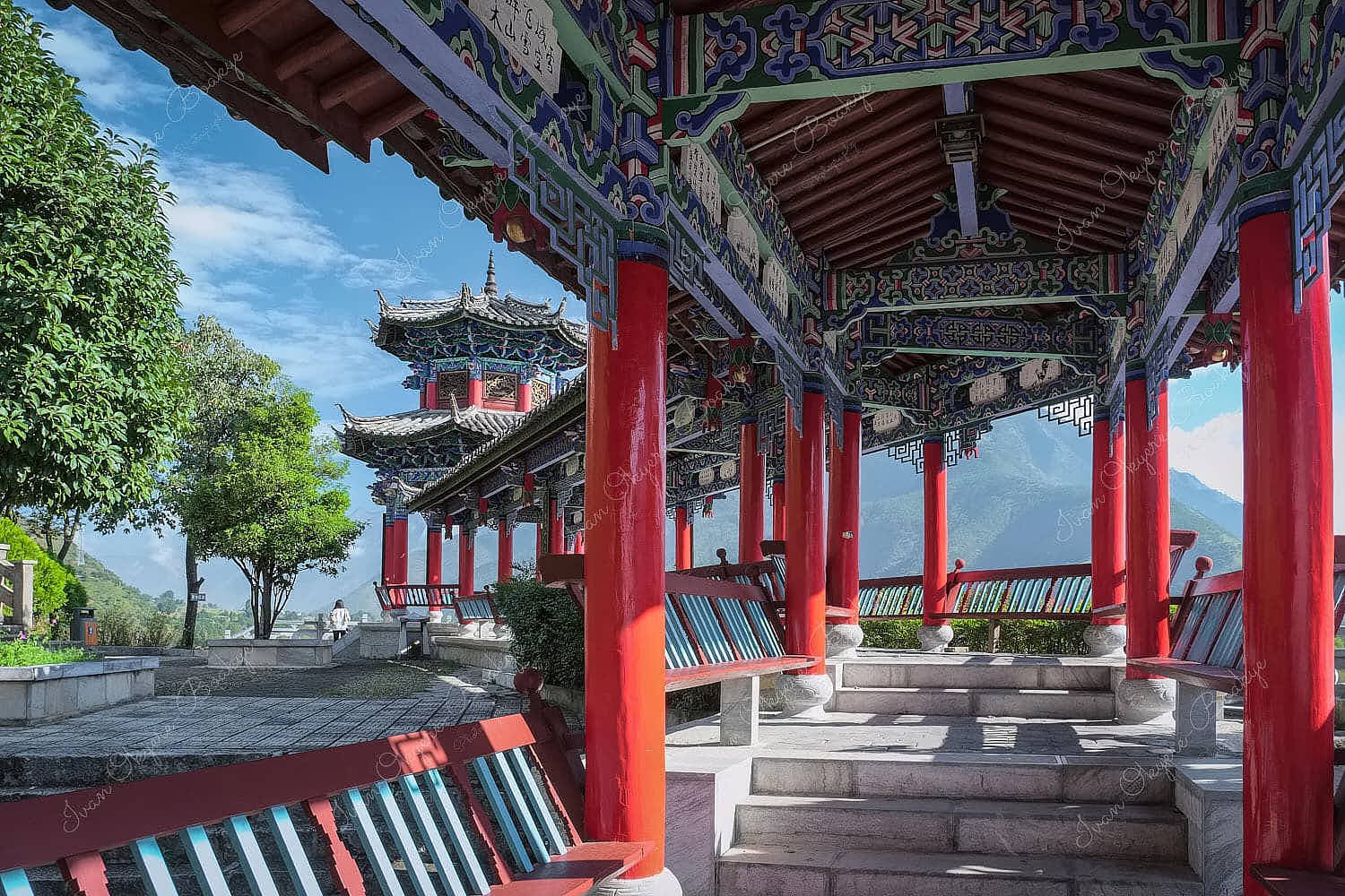 A red and blue coloured Chinese design style covered walkway with rest benches leading to a panoramic viewpoint in Stone Drum Town, Shangri-La, China